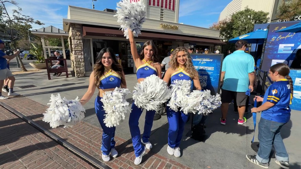 Three cheerleaders in blue and yellow uniforms holding white pom-poms pose for the camera outside a store, with a crowd and signage in the background.