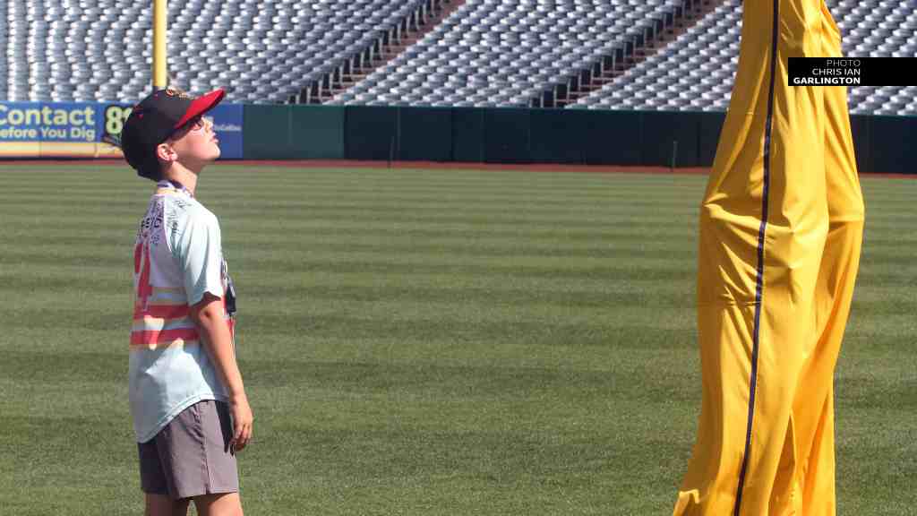 A young boy in a cap and sunglasses looks up at a tall, yellow fabric structure on a baseball field, surrounded by empty seating in the background.