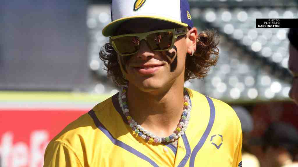A young man with curly hair wearing a yellow sports jersey and sunglasses, smiling and standing in an outdoor setting.