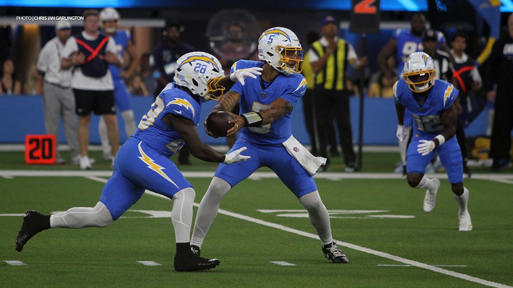 A professional football player in blue jersey #5 prepares to receive the ball from another player in blue jersey #28 during a game. Two additional players in blue are positioned in the background.